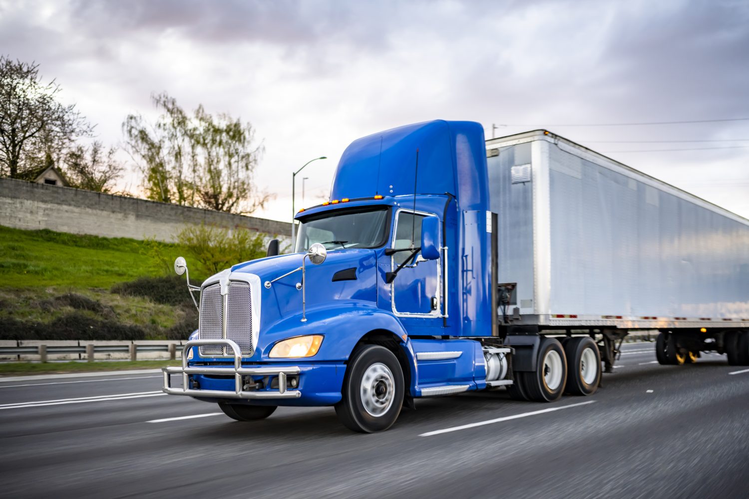 Day cab blue big rig industrial semi trucks tractor with roof spoiler and turn on headlight transporting commercial cargo in dry van semi trailer running on the wide highway road at twilight time