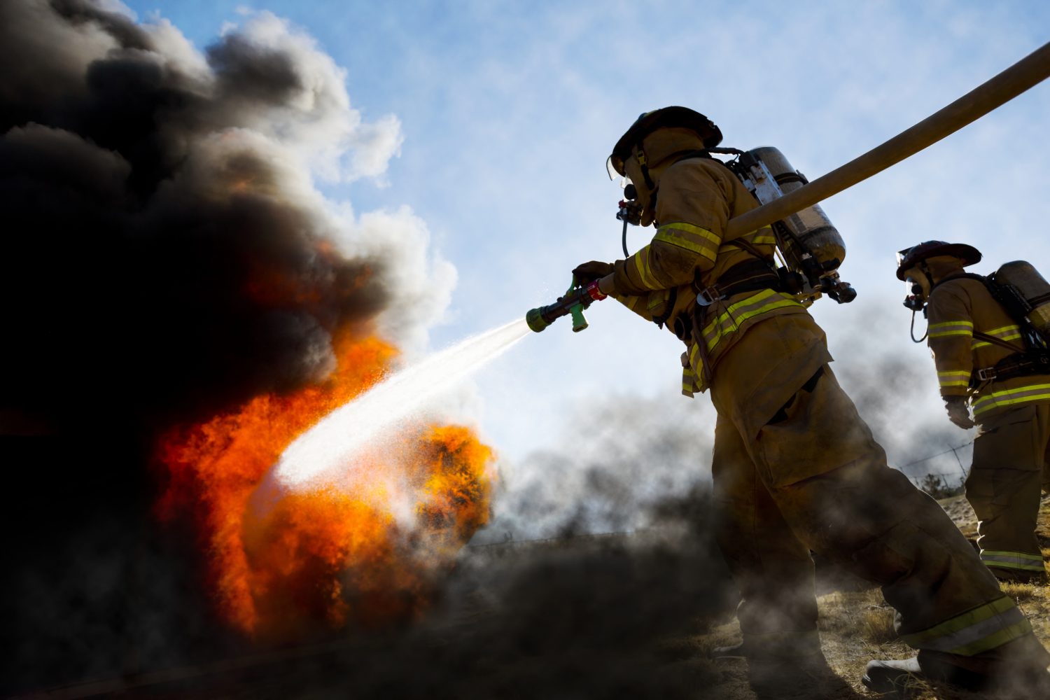 Firefighters in a fire protection suit wearing firefighter helmet with breathing device and holding fire hose is extinguishing a burning house fire that is putting off excessive heat and smoke.  Fire could have been caused by accident or arson.  Second firefighter in background.