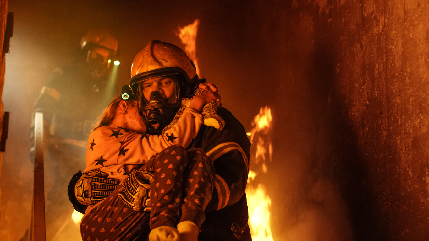 Brave Fireman Descends Stairs of a Burning Building and Holds Saved Girl in His Arms. Open fire and one Firefighter in the Background.